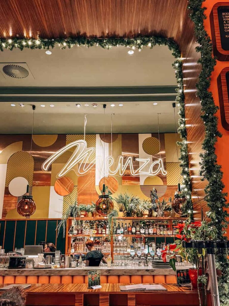 Interior of Menza restaurant in Budapest with a stylish bar area, warm lighting, greenery, hanging pendant lights, and a neon Menza sign creating a cozy modern atmosphere.