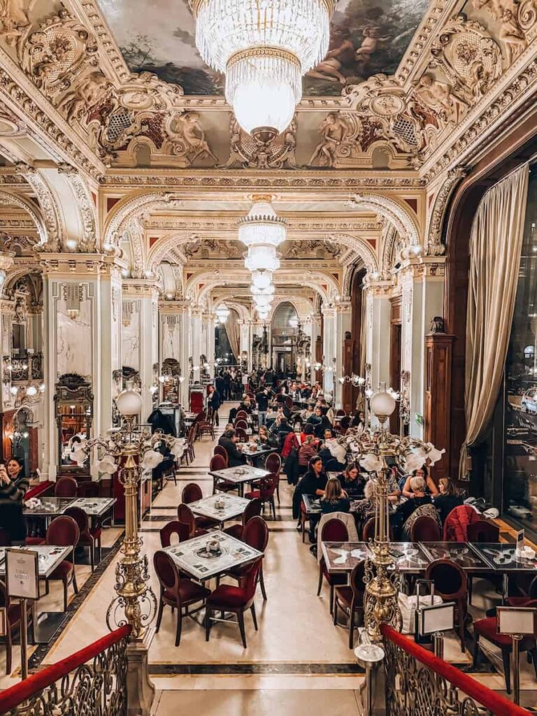 Opulent interior of New York Café in Budapest with gilded columns, ornate arches, crystal chandeliers, and rows of red velvet chairs filled with guests dining in the historic café.