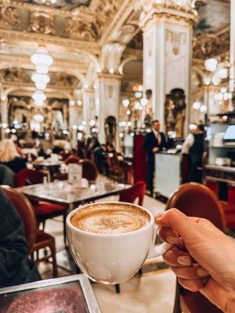 Hand holding a cup of coffee inside New York Café in Budapest with the grand gilded interior softly blurred in the background, showcasing chandeliers, marble tables, and the elegant café atmosphere.
