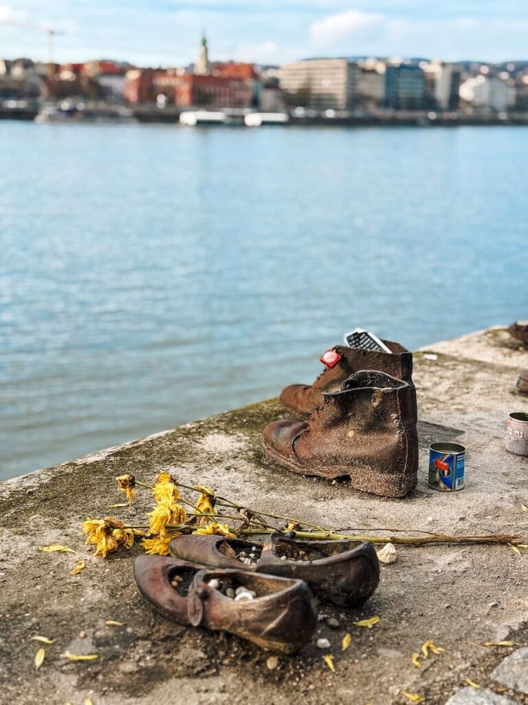 Bronze shoes arranged along the edge of the Danube River in Budapest as part of the Shoes on the Danube Bank memorial, with weathered footwear, flowers placed in remembrance, and the river flowing quietly in the background.