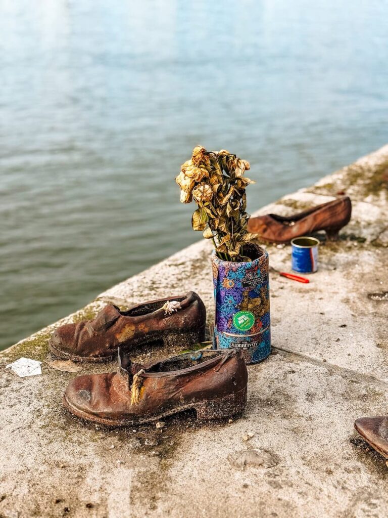 Close view of rusted bronze shoes at the Shoes on the Danube Bank memorial in Budapest, accompanied by dried flowers in a tin can, set on the riverbank with the Danube water stretching behind them in a solemn and reflective scene.