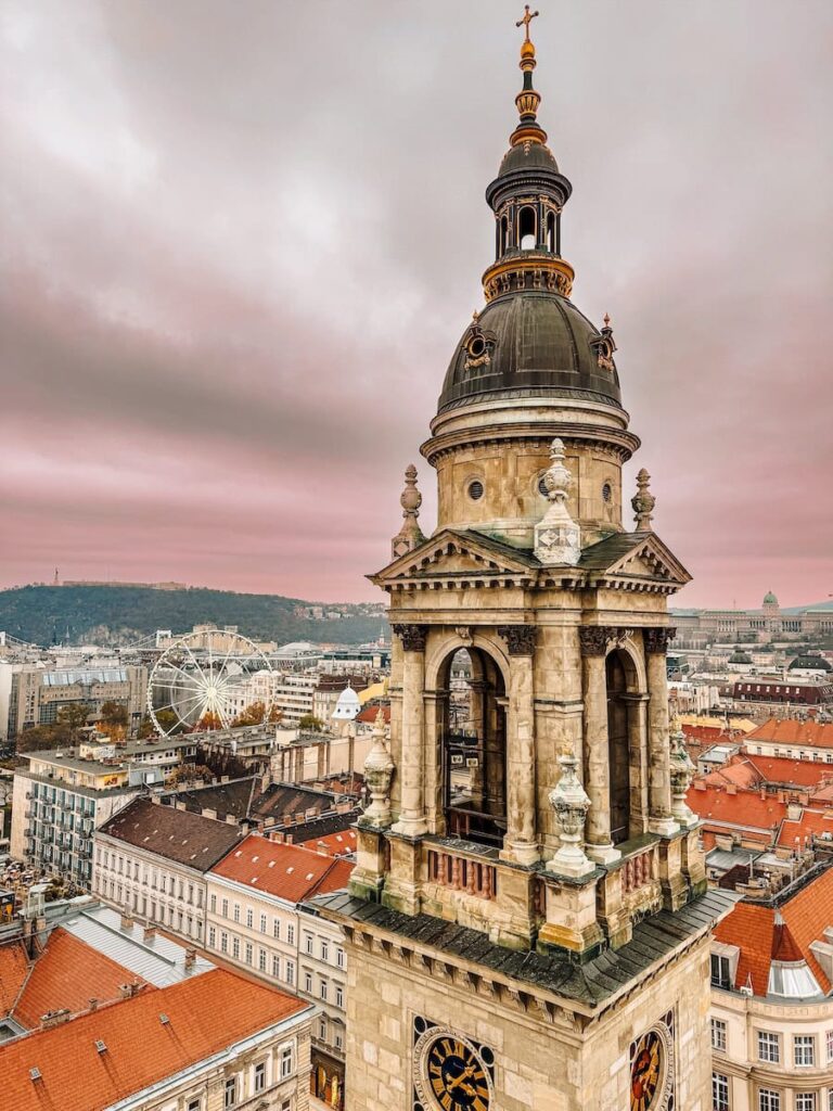 View from the dome terrace of St Stephen’s Basilica looking out over Budapest with the bell tower in the foreground, red tiled rooftops below, a Ferris wheel in the distance, and a soft pink cloudy sky.