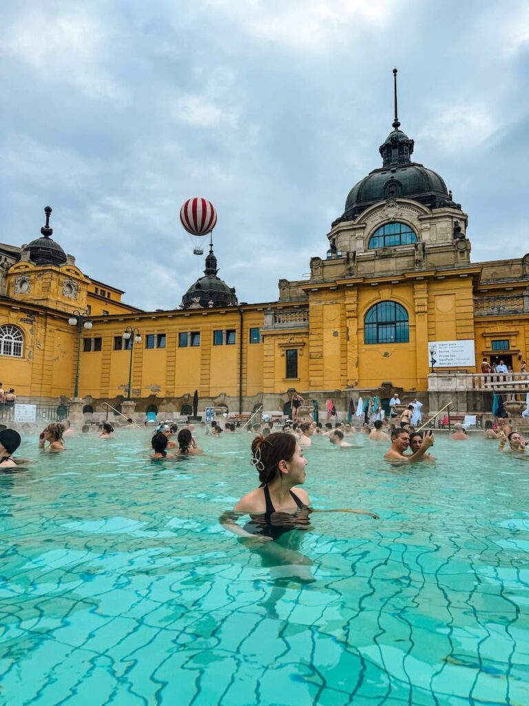 Outdoor pools at the Széchenyi Thermal Bath in Budapest filled with people soaking in bright turquoise water, surrounded by the grand yellow neo Baroque bathhouse with domed rooftops under a cloudy sky.