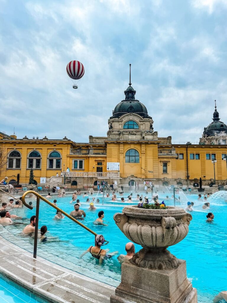 Crowded open air thermal pool at Széchenyi Baths in Budapest with steaming blue water, bathers relaxing along the pool edges, and the iconic yellow historic building and domes rising in the background.
