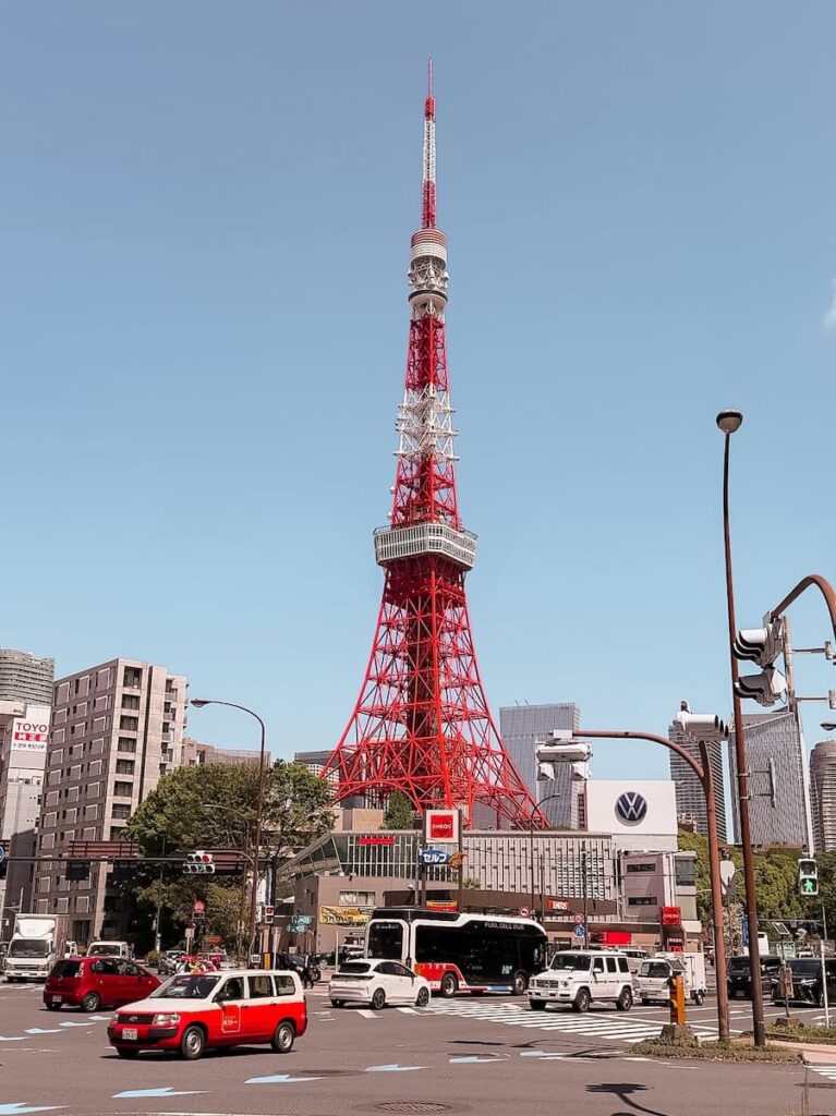 Tokyo Tower towering over a busy city intersection with cars buses and modern buildings under a clear blue sky