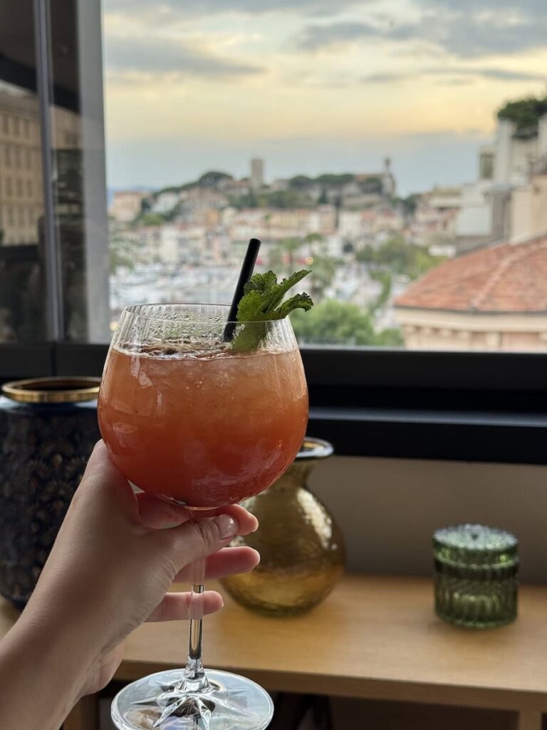 Hand holding a cocktail at Le Roof restaurant with panoramic views over Cannes and Le Suquet in the background