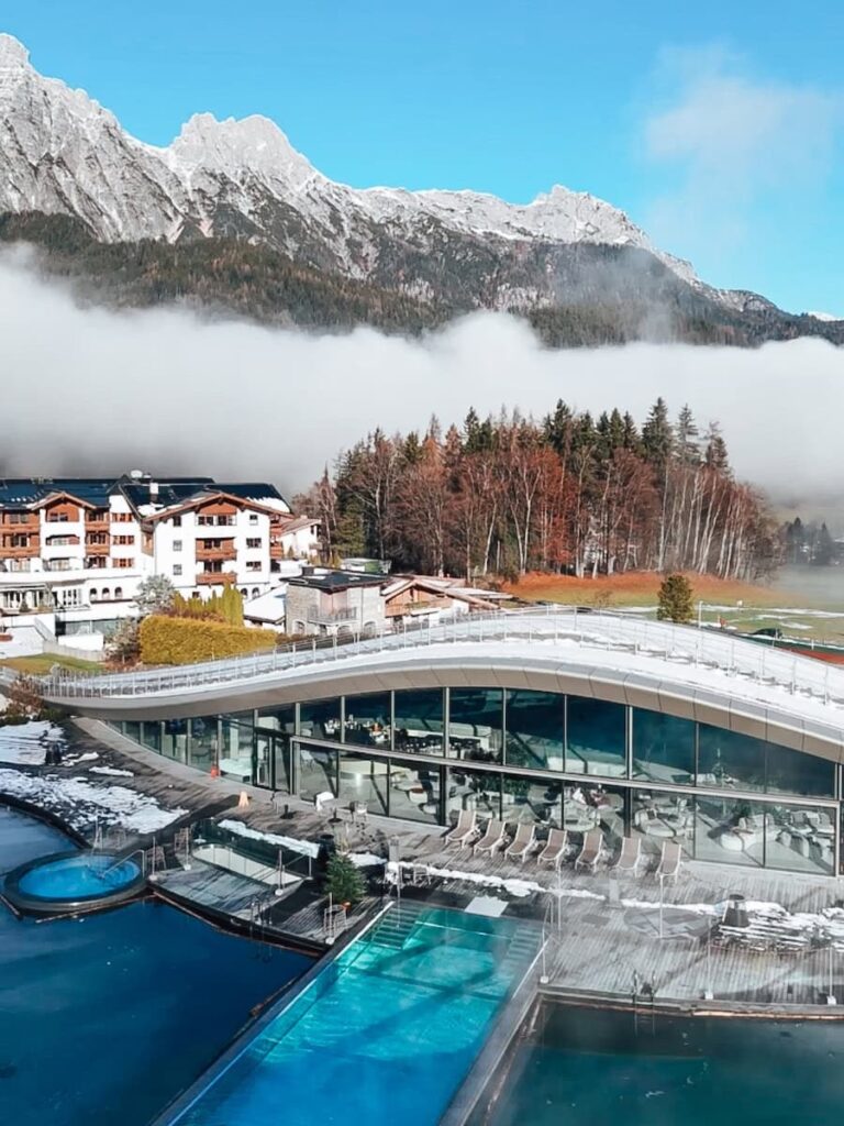 Aerial view of Hotel Krallerhof spa complex with outdoor pools modern architecture and alpine mountains rising above low clouds