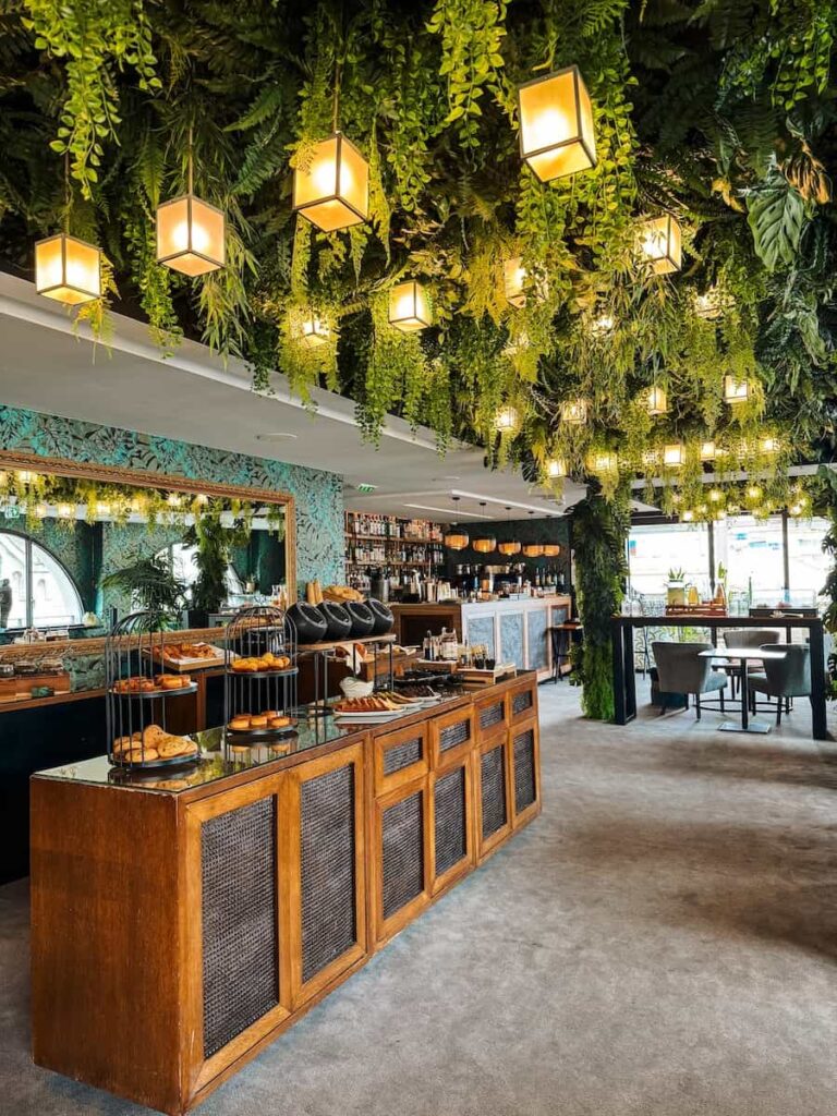 Breakfast buffet at Five Seas Hotel Cannes with a lush green ceiling, warm hanging lights, wooden buffet counters, and elegant dining area filled with natural light