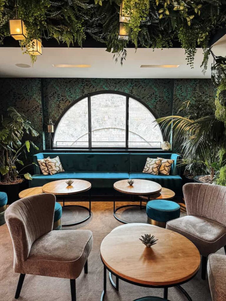 Cozy dining lounge at Five Seas Hotel Cannes with teal velvet seating, round wooden tables, lush greenery, and a large arched window creating a calm boutique atmosphere