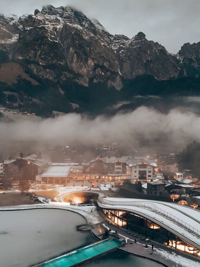 Aerial view of Hotel Krallerhof in winter surrounded by snowy pools glowing lights and dramatic alpine mountains