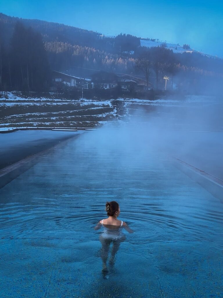 Woman relaxing in an outdoor thermal pool at Hotel Krallerhof with steam rising and forested hills behind