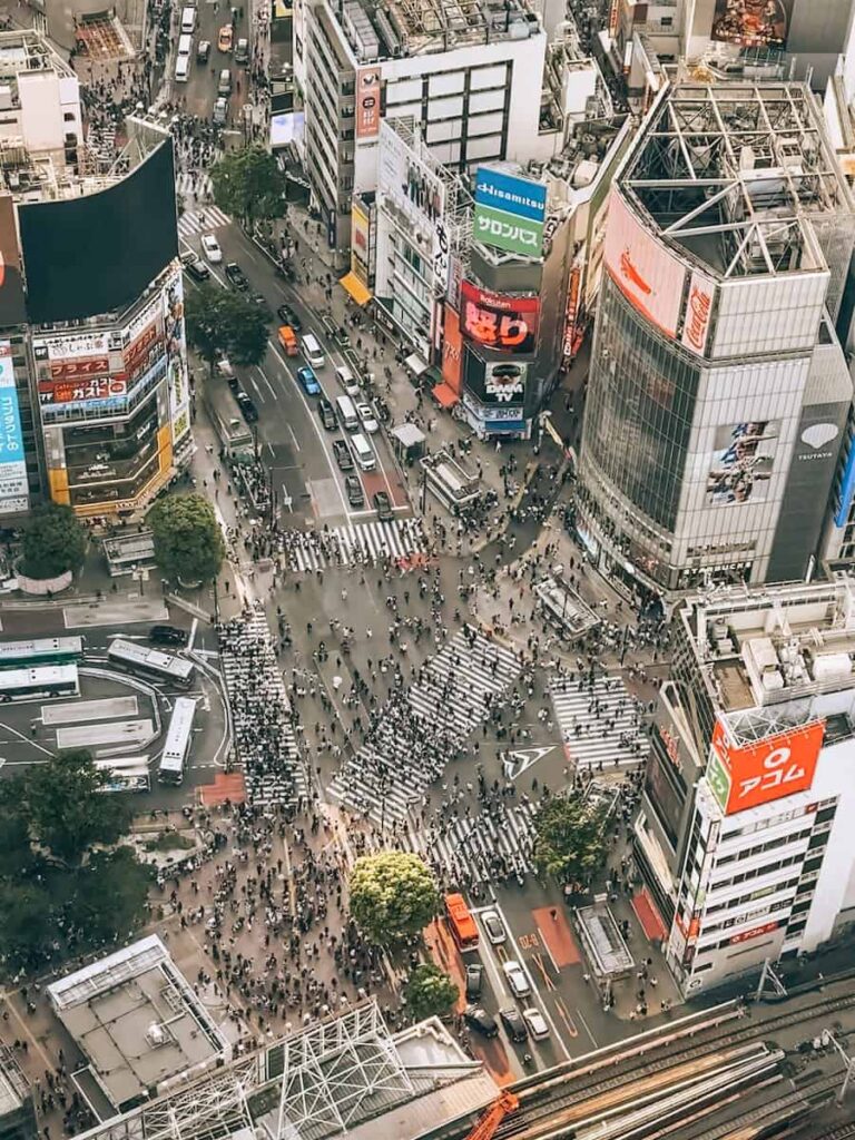 Aerial view of Shibuya Scramble Crossing showing streams of pedestrians crossing in multiple directions with large billboards and city streets below
