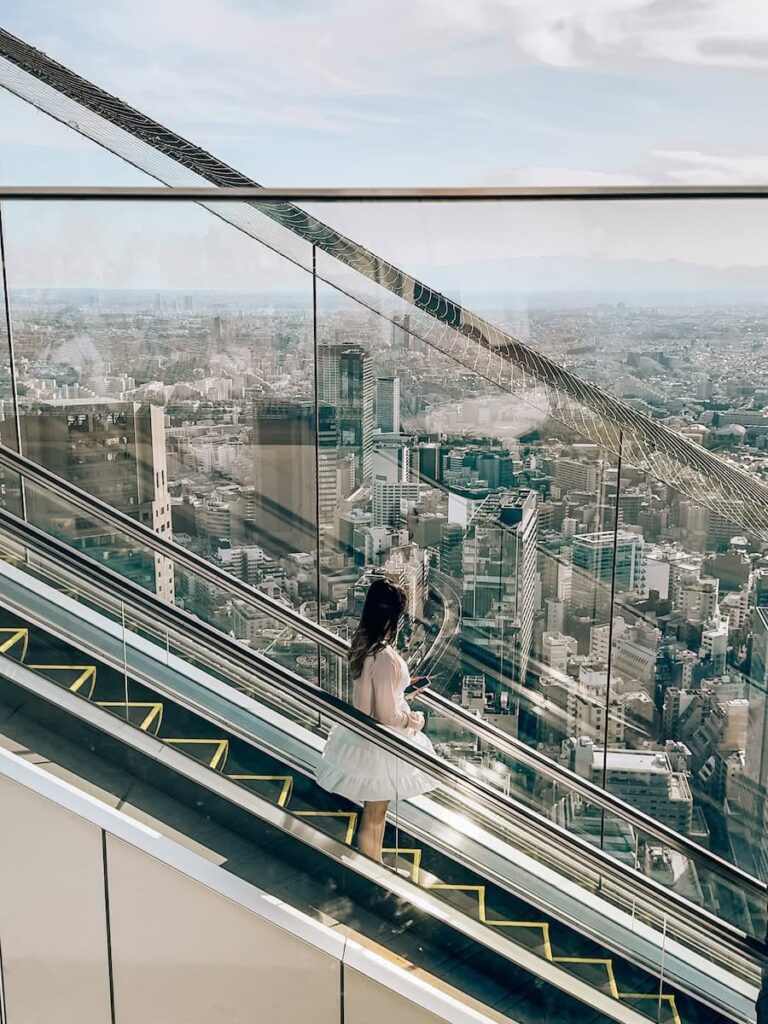 Woman riding an escalator at Shibuya Sky observation deck with panoramic views of Tokyo city stretching into the distance