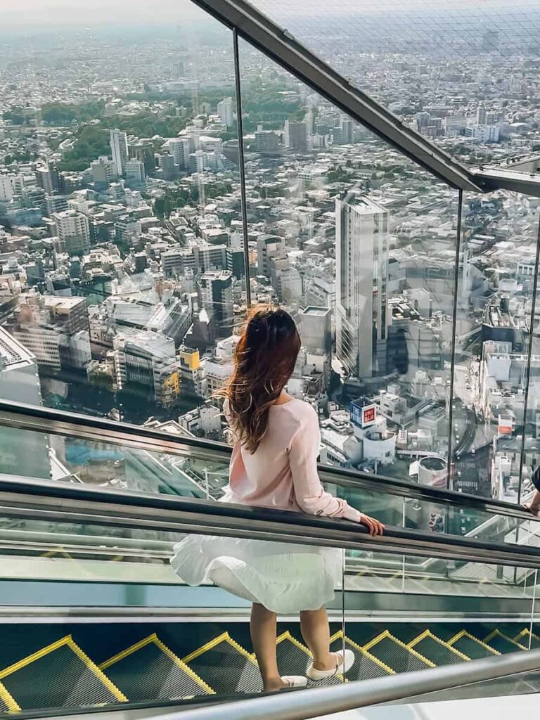 Author standing on an escalator inside Shibuya Sky with glass walls and sweeping views of the Tokyo skyline below