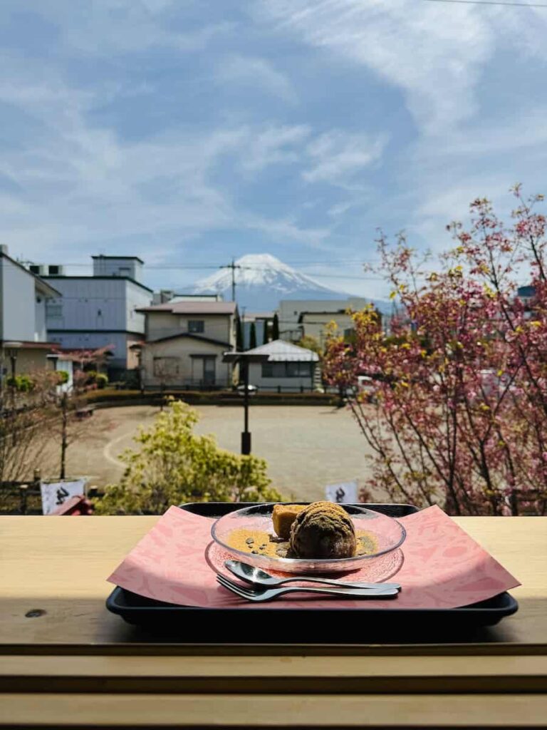 Dessert on a wooden table inside a cafe in Shimoyoshida with Mount Fuji visible through the window and cherry blossoms outside on a clear day