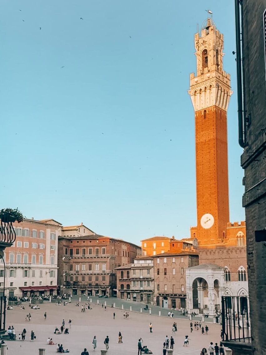 Piazza del Campo in Siena, Italy, with people scattered across the spacious square, surrounded by historic buildings, and the tall Torre del Mangia tower rising prominently against a clear blue sky.
