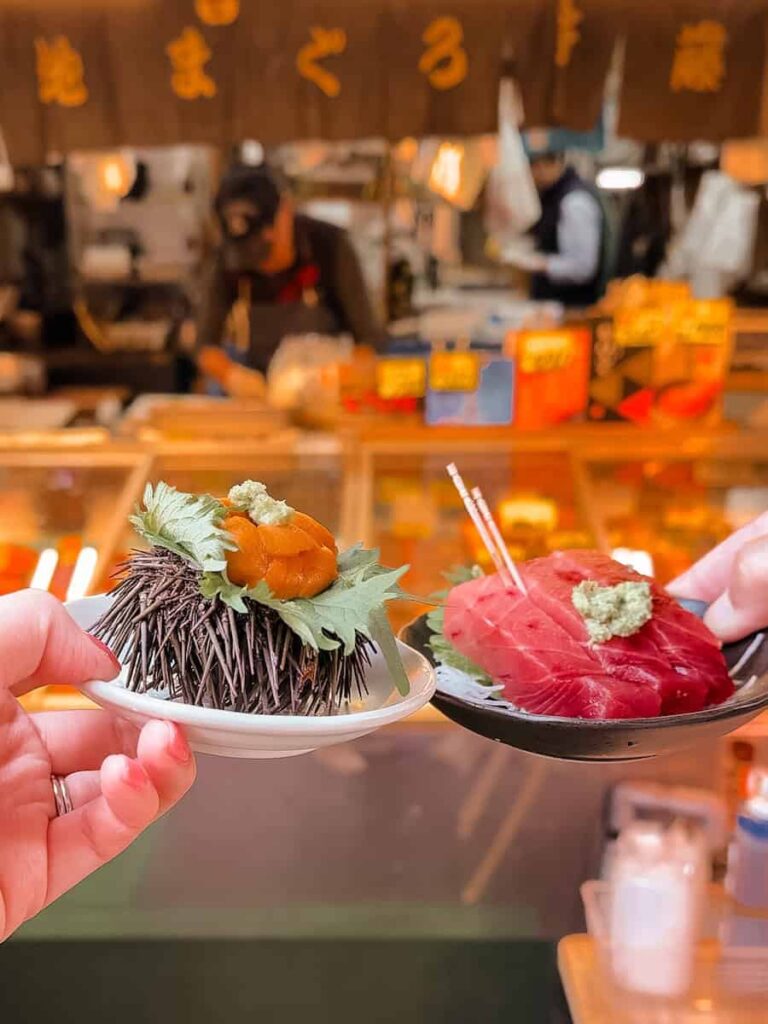 Hand holding fresh uni and tuna sashimi on small plates at Tsukiji Outer Market in Tokyo with a seafood stall in the background
