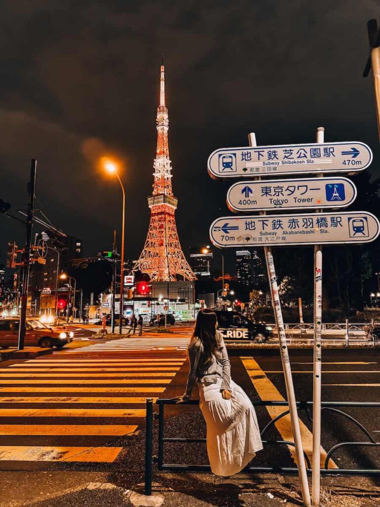 The author near a crosswalk at night with Tokyo Tower illuminated in the background and city street signs pointing toward the landmark