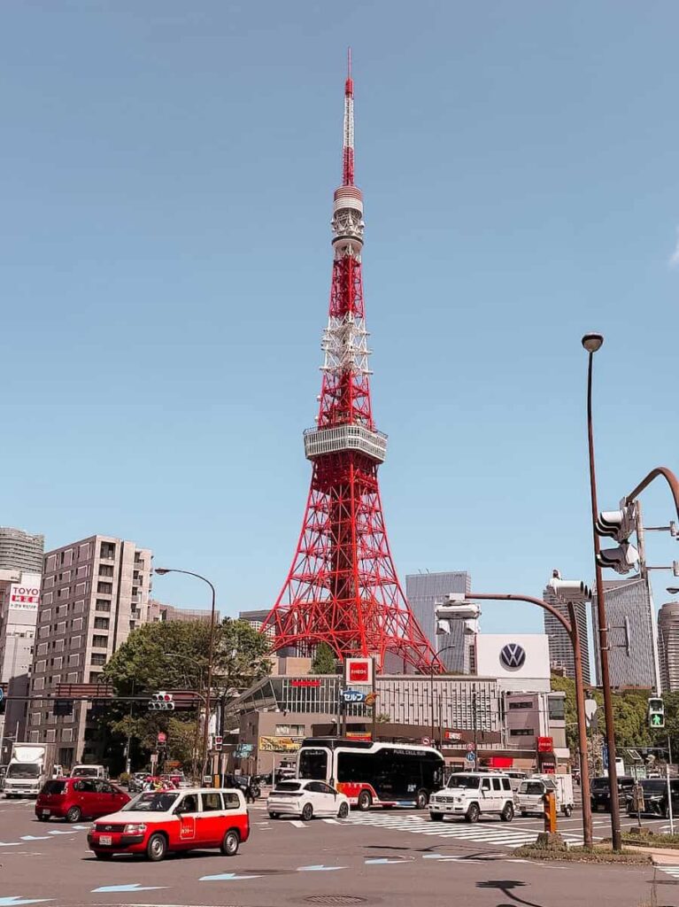 Tokyo Tower rising above a busy city intersection in Tokyo during the day with cars buses and surrounding buildings under a clear blue sky