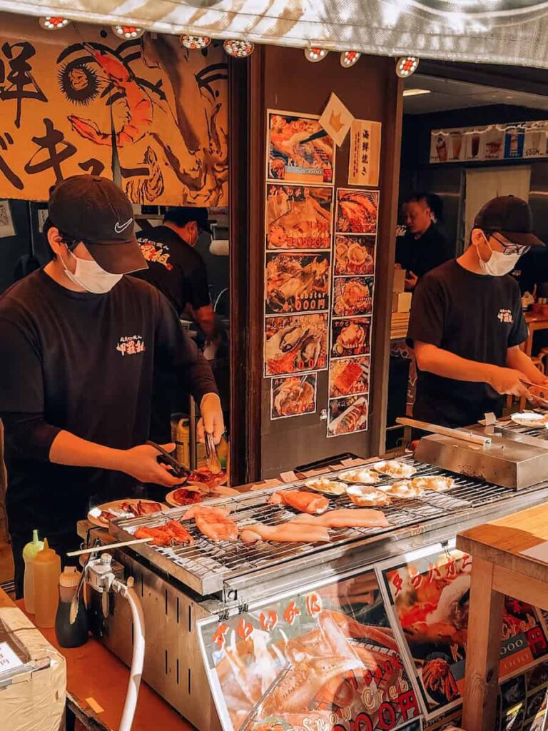Seafood vendors grilling fresh fish and shellfish at Tsukiji Outer Market in Tokyo with menu signs and a busy food stall atmosphere