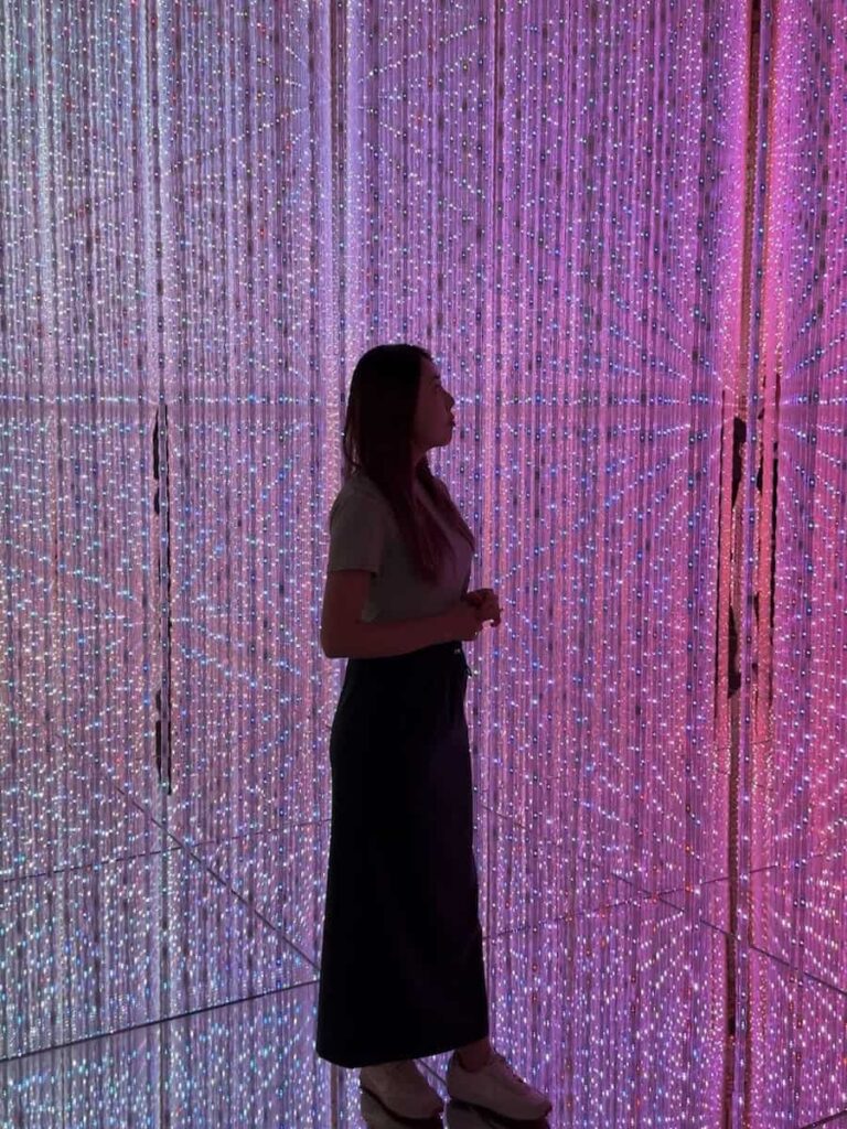 Woman standing inside a glowing immersive light installation at teamLab Borderless in Tokyo surrounded by mirrored walls and pink and purple LED lights