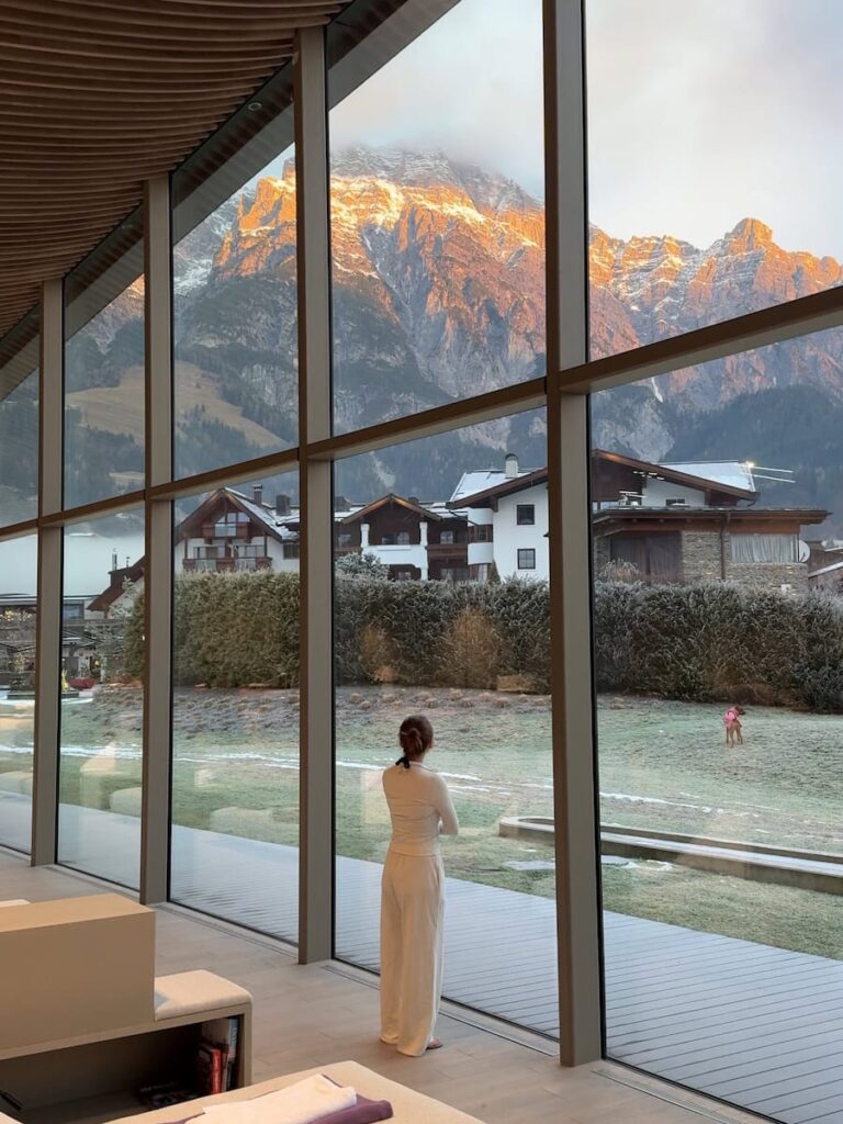 Woman standing in a relaxation room at Hotel Krallerhof looking out through floor to ceiling windows toward alpine houses and sunlit mountains