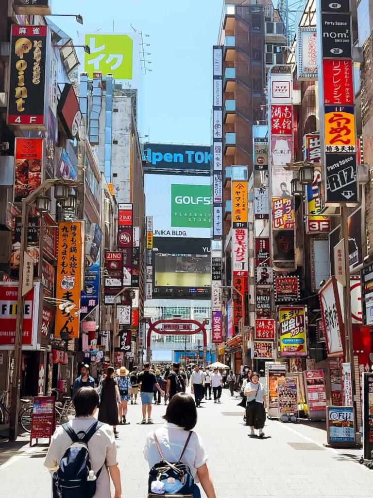 Busy Tokyo street in Shinjuku during the day with colorful vertical Japanese shop signs people walking through the entertainment district and bright urban city scenery. If you're wondering where to stay in Tokyo, shinjuku is the best
