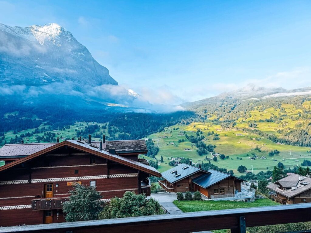 View from a balcony in Grindelwald, Switzerland overlooking traditional wooden chalets, green alpine hills, low clouds, and snow capped mountains in the distance.