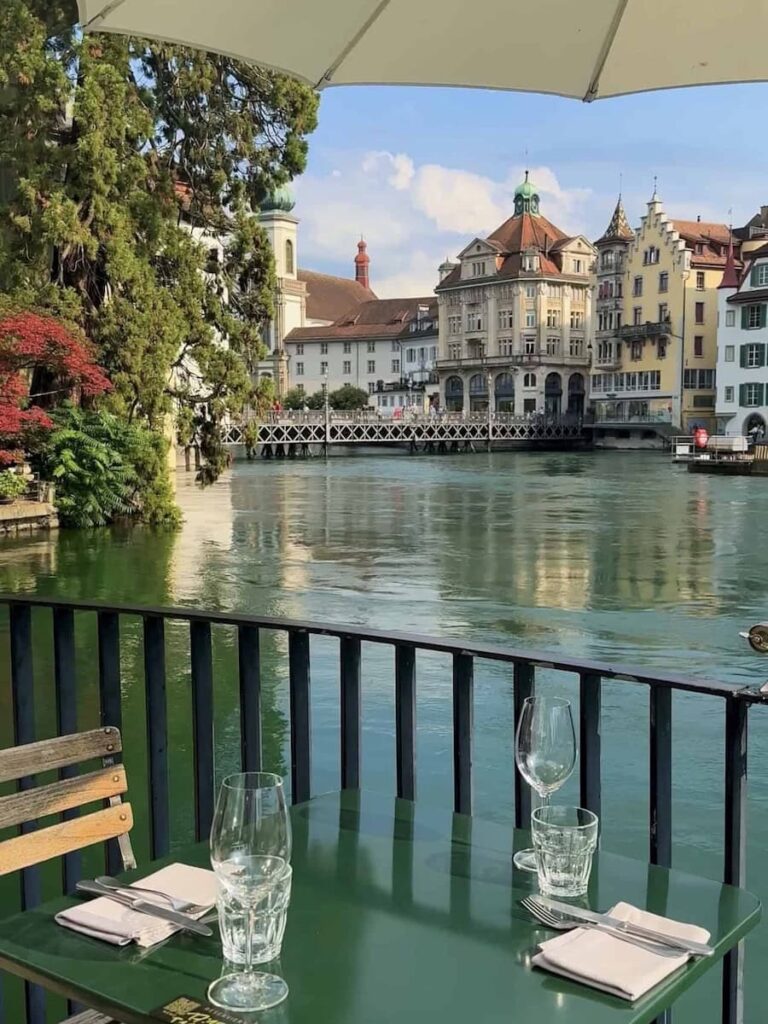Riverside outdoor dining table in Lucerne, Switzerland overlooking turquoise water, historic buildings, and the famous Chapel Bridge area on a sunny day.