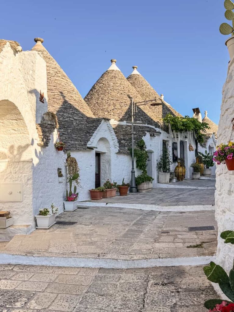 Traditional trulli houses lining a quiet street in Alberobello, with whitewashed stone walls, iconic conical roofs, colorful flower pots, and lush climbing vines.