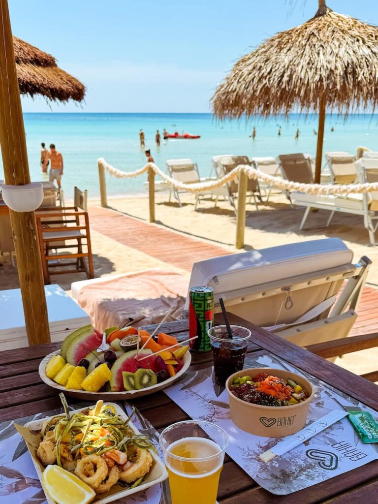 Lunch overlooking the beach at Bahia Porto Cesareo Beach Club, with fresh seafood, tropical fruit, turquoise water, sun loungers, and thatched umbrellas along the Ionian coast of Puglia.