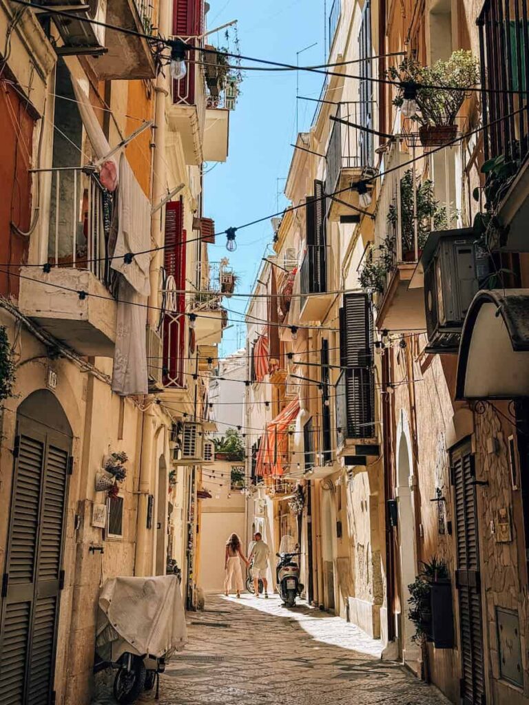 A narrow stone alley in Bari Vecchia lined with colorful shutters, balconies, hanging lights, and potted plants, with a couple walking hand in hand through the historic old town.
