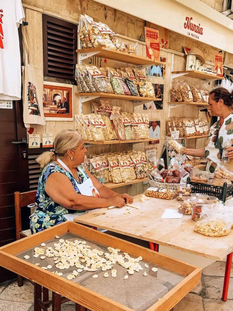 Traditional orecchiette pasta being hand rolled by a local woman on Strada delle Orecchiette in Bari Vecchia, with fresh pasta drying on trays and bags of handmade pasta displayed outside the shop.