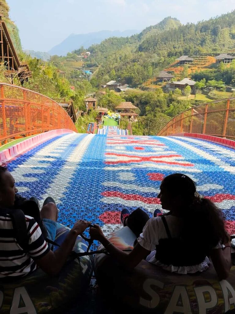 Visitors preparing to ride the rainbow slide at Cat Cat Village near Sapa, Vietnam, overlooking the surrounding hills and traditional village landscape.