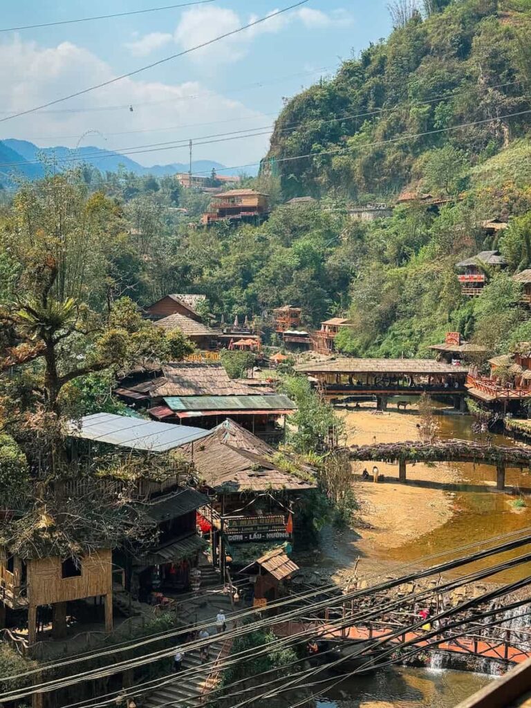 Panoramic view of Cat Cat Village near Sapa, Vietnam, with traditional wooden buildings, bridges, and a stream winding through the valley.