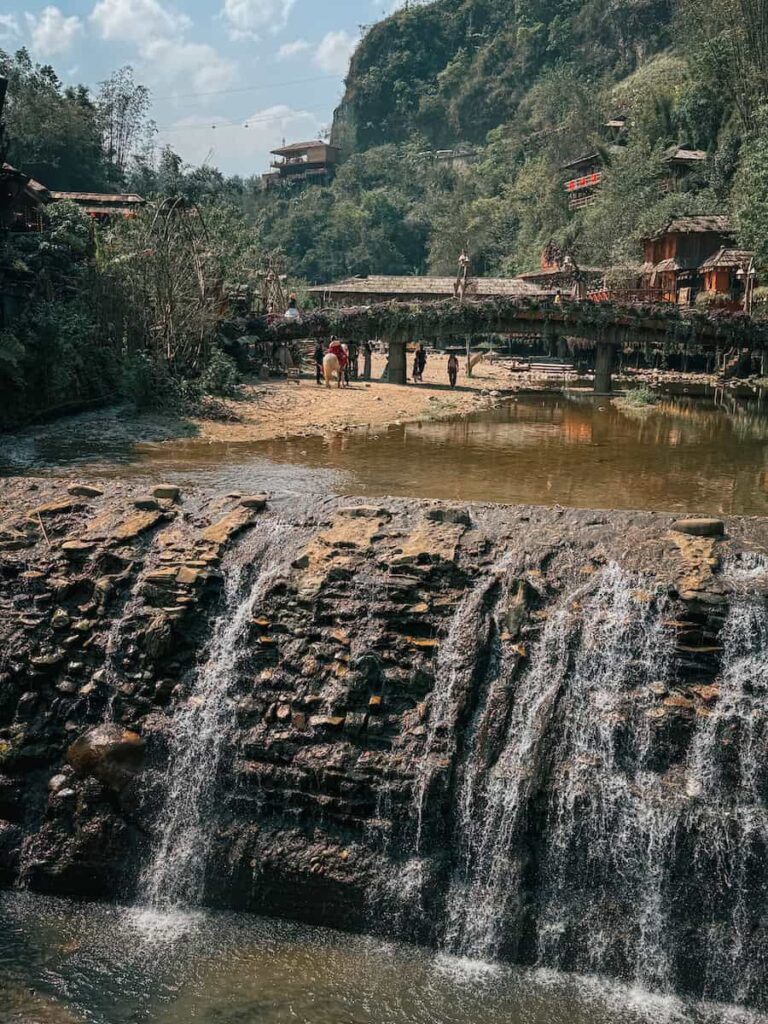 Waterfall flowing through Cat Cat Village near Sapa, Vietnam, with a pedestrian bridge and traditional village buildings in the background.