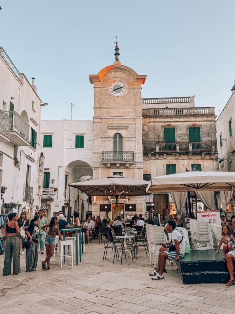 Historic clock tower overlooking Piazza Vittorio Emanuele in Cisternino, with outdoor cafés, limestone buildings, and people enjoying the lively town square at sunset.
