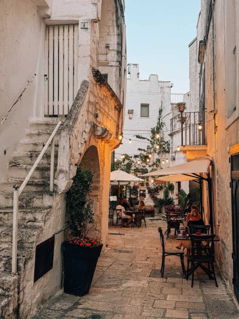 Narrow stone alley in the historic center of Cisternino lined with whitewashed buildings, outdoor café tables, flower pots, and string lights in Puglia, Italy.