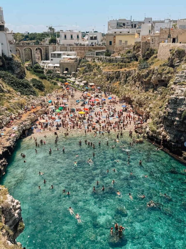 Panoramic view of Lama Monachile Beach in Polignano a Mare, where swimmers enjoy crystal clear turquoise water between towering limestone cliffs beneath the historic old town.