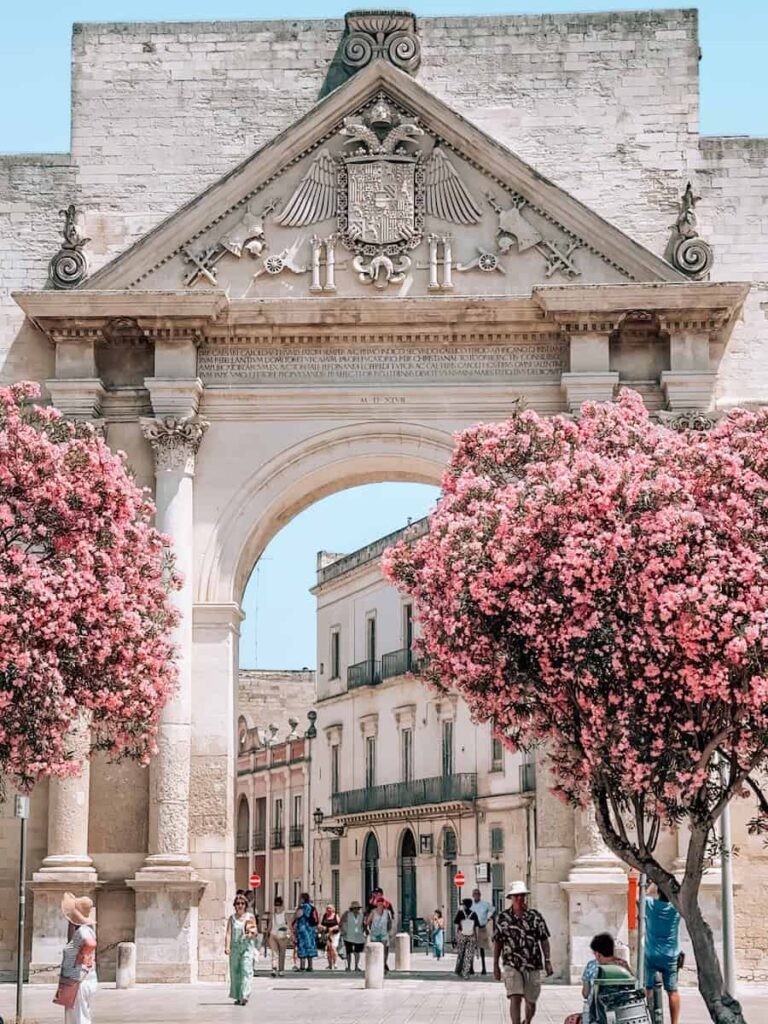Porta Napoli, the triumphal gateway marking the entrance to Lecce's historic center, framed by blooming pink oleander trees on a sunny summer day.