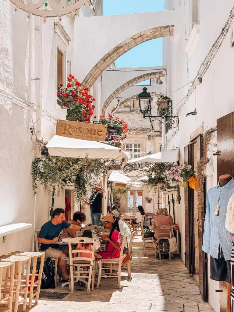 Outdoor cafés lining a charming whitewashed alley in Locorotondo, with flowering balconies, stone archways, and traditional limestone buildings in the historic center.