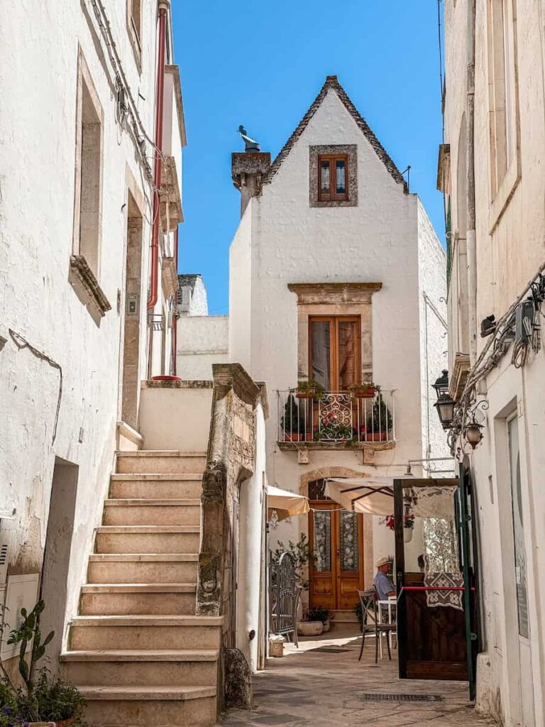 Quiet whitewashed alley in Locorotondo featuring traditional limestone buildings, an exterior stone staircase, and a historic house with a flower filled balcony in Puglia, Italy.