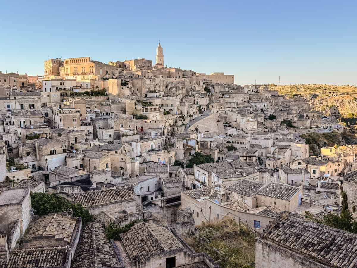 Panoramic view of Matera, Italy, showing the ancient Sassi districts with hundreds of pale limestone houses and cave dwellings cascading down the hillside. The Matera Cathedral rises above the city on the ridge under a clear blue sky, illuminated by warm golden hour light.