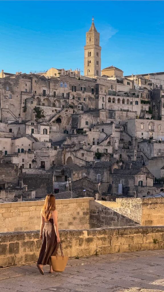 A woman walking and overlooking the Sassi district in Matera, Italy, with the ancient limestone cave dwellings and the Matera Cathedral illuminated by warm sunrise light beneath a clear blue sky.