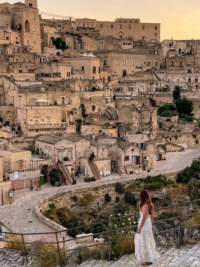 Woman in a white dress overlooking the Sassi di Matera at sunset, with the ancient limestone cave dwellings and historic stone buildings cascading down the hillside.