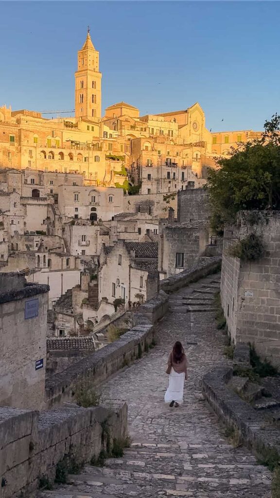 A woman walks along a historic stone lane in the Sassi district of Matera, Italy, toward the Matera Cathedral as the ancient limestone buildings glow in warm sunrise light.