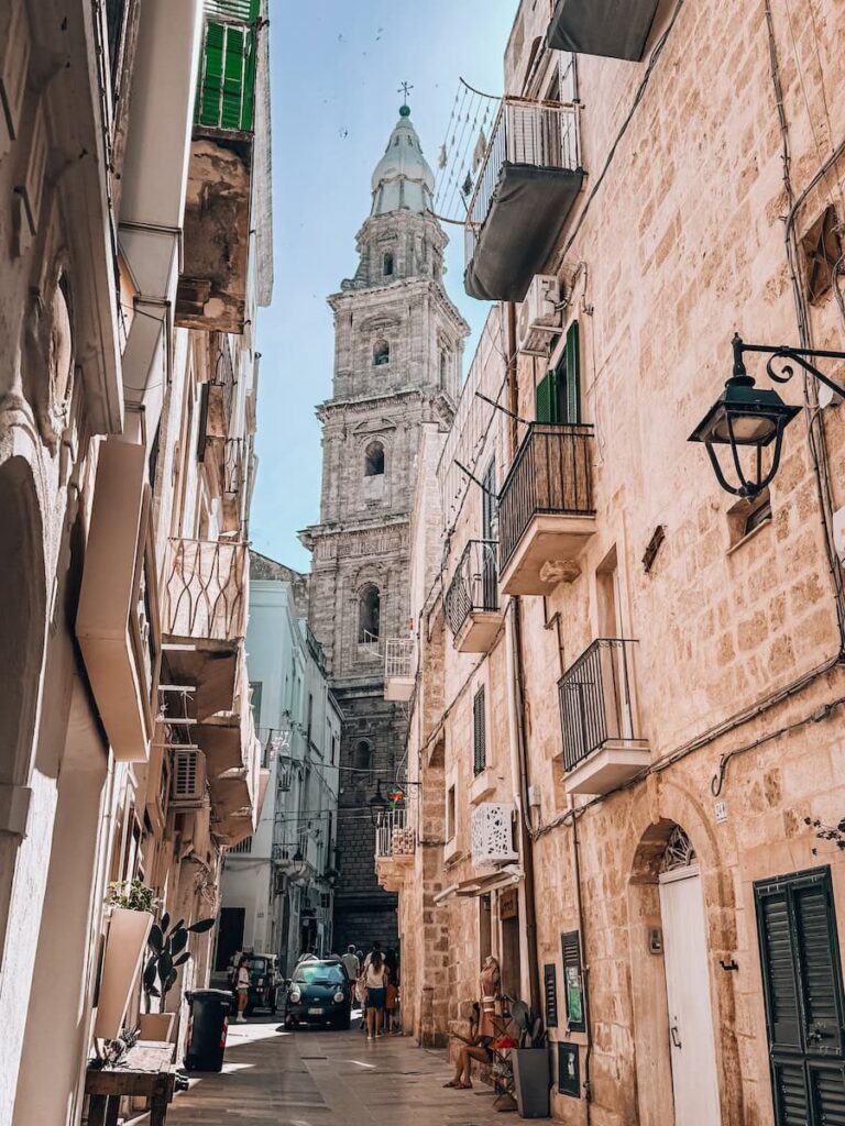 Historic street in Monopoli lined with limestone buildings and balconies, leading toward the bell tower of the Cathedral of Maria Santissima della Madia in Puglia, Italy.