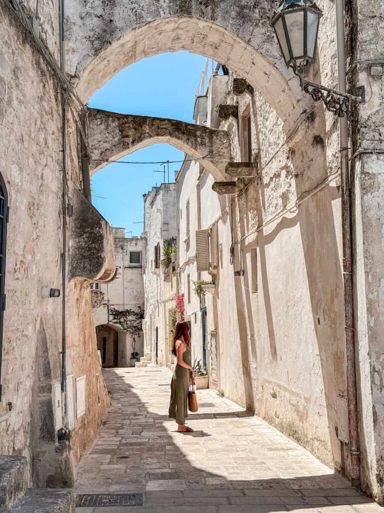Woman walking beneath historic stone arches along a quiet whitewashed alley in Ostuni's historic center, known as the White City of Puglia.