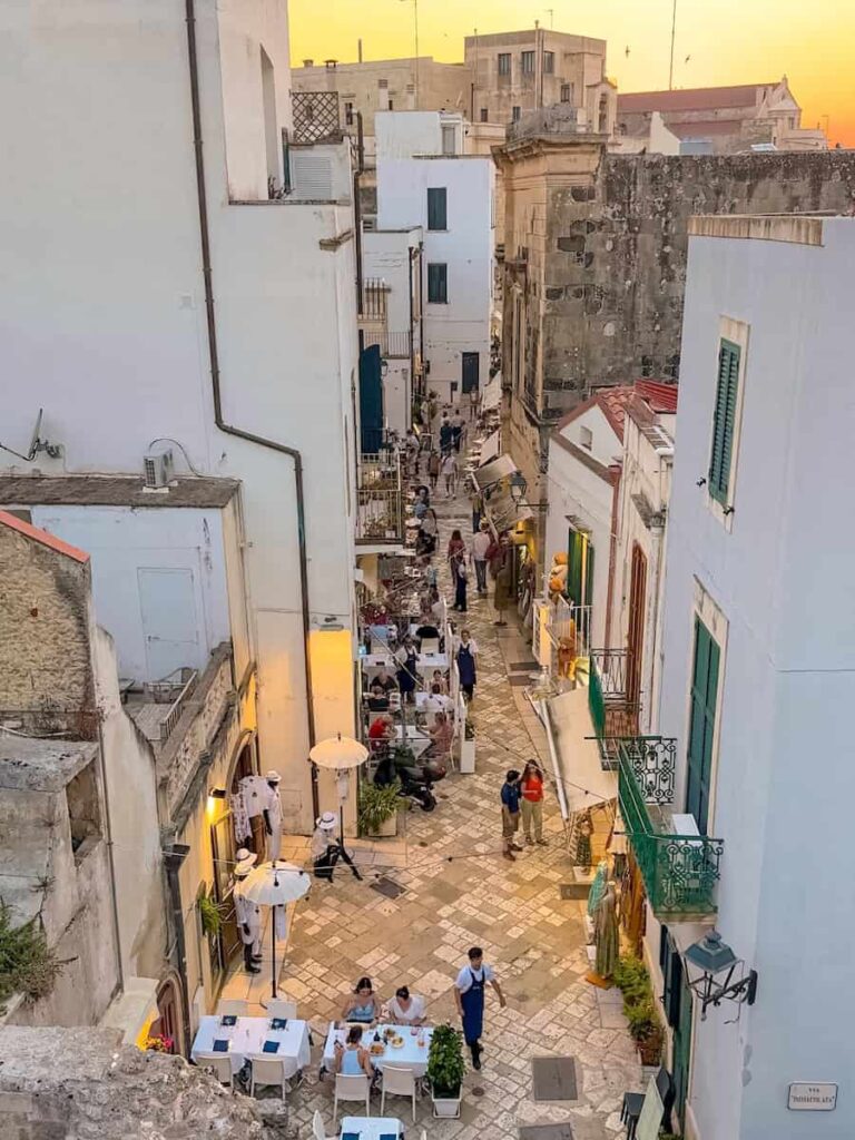 View down a lively pedestrian street in Otranto's historic center at sunset, lined with whitewashed buildings, outdoor restaurants, boutiques, and colorful balconies.