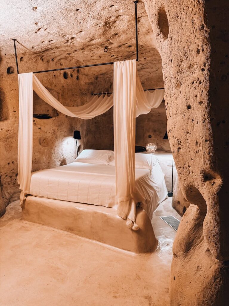Cave suite bedroom at Sant'Angelo Luxury Resort in Matera, Italy, featuring a white canopy bed carved into a historic limestone cave with natural rock walls, warm ambient lighting, and minimalist furnishings.